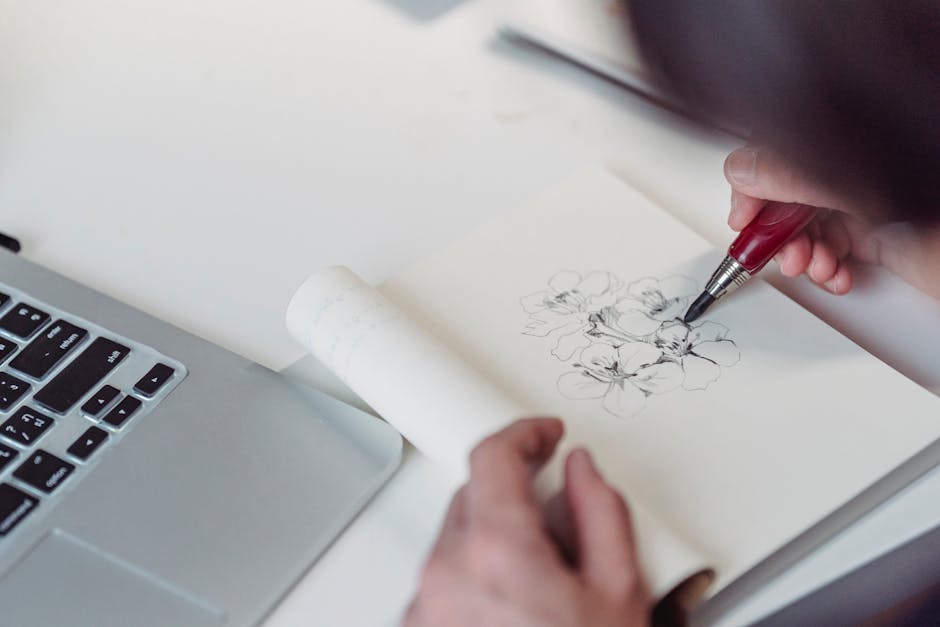 Close-up of a person sketching flowers with a pen next to a laptop, combining creativity and technology.