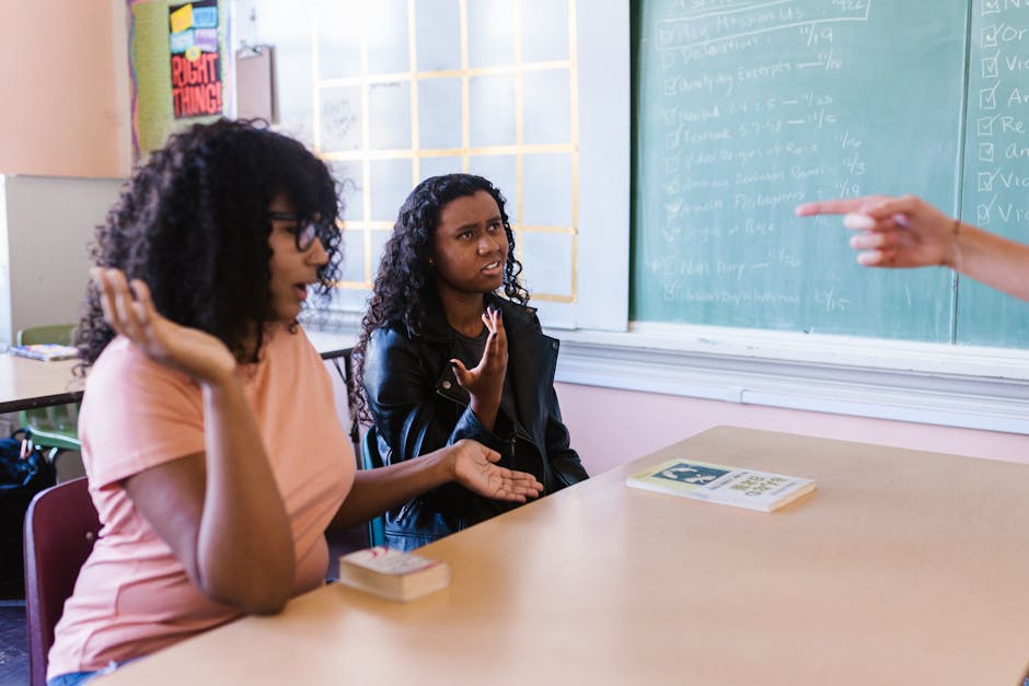 Two students in a classroom discussing content with a teacher pointing at a blackboard.