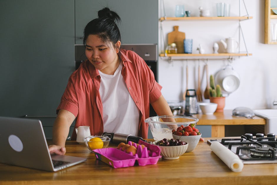 A young adult using a laptop while preparing food in a modern kitchen setting.