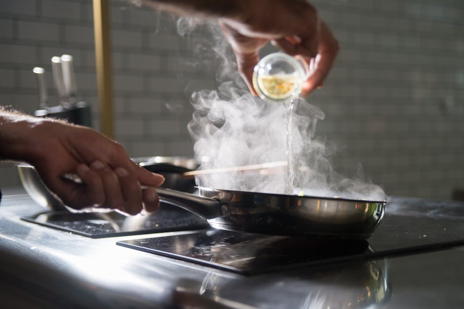 Close-up of a chef adding liquid to a steaming frying pan in a modern kitchen.