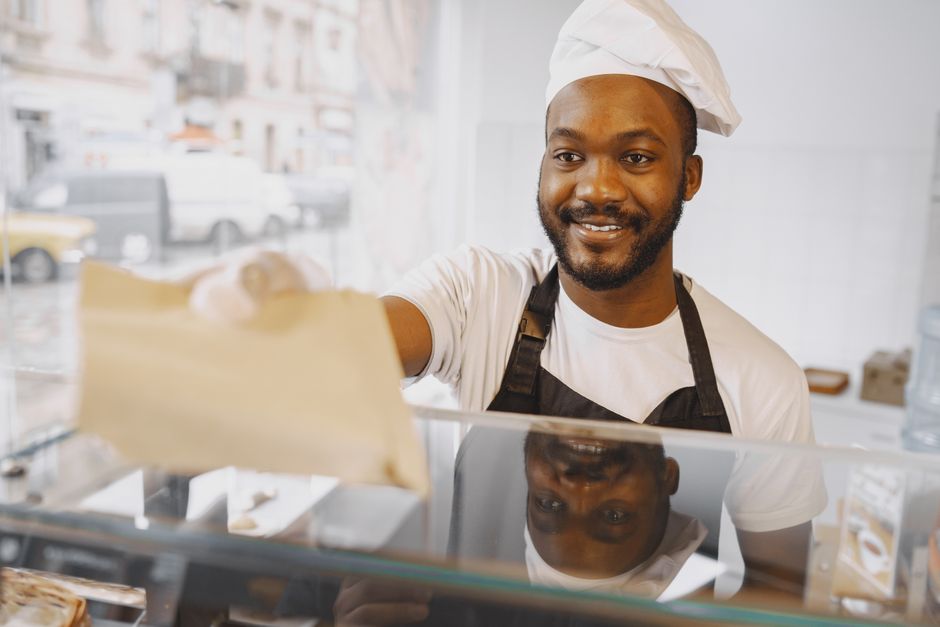 Cheerful baker in apron and toque blanche offers a baked good in a paper bag from a bakery counter.
