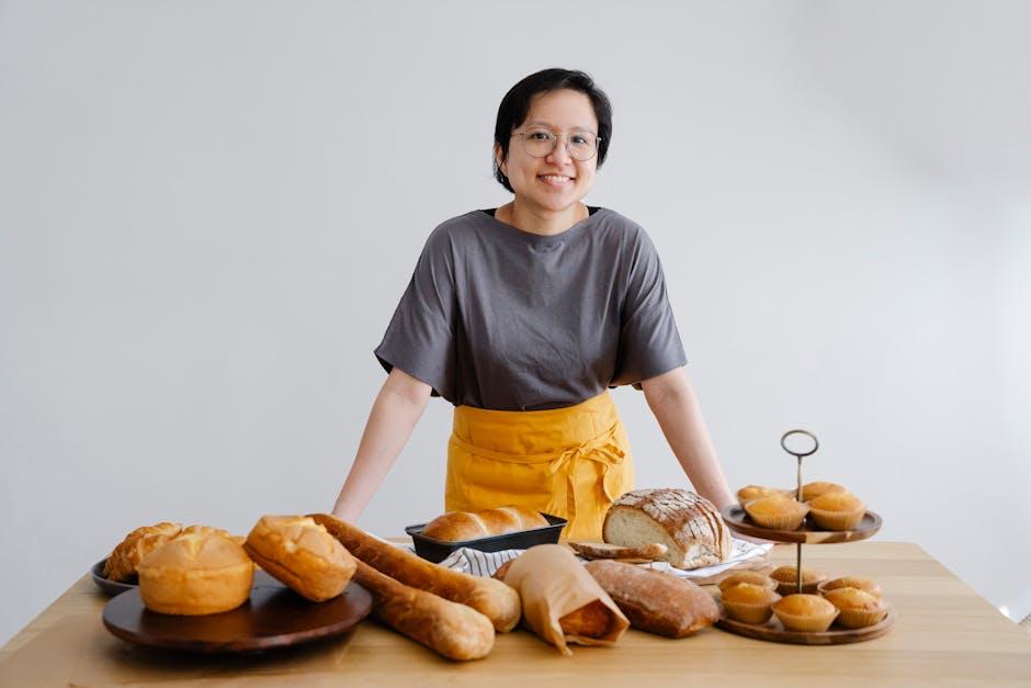Smiling Asian female baker showcasing a variety of fresh bread and pastries indoors.