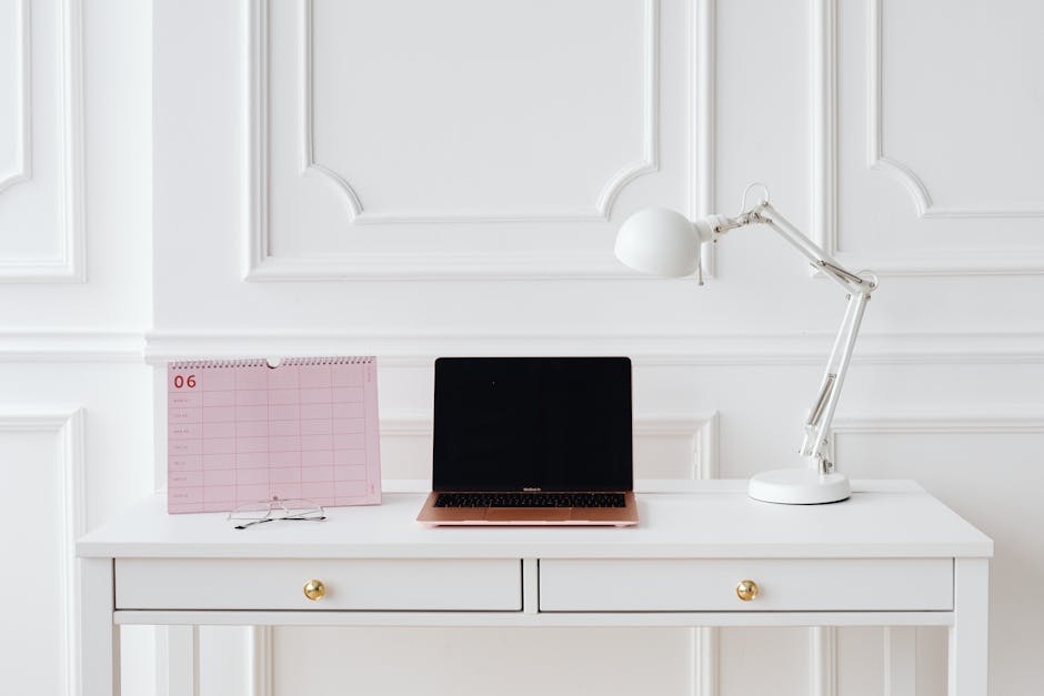 A minimalist workspace featuring a laptop, desk lamp, and calendar on a white desk.