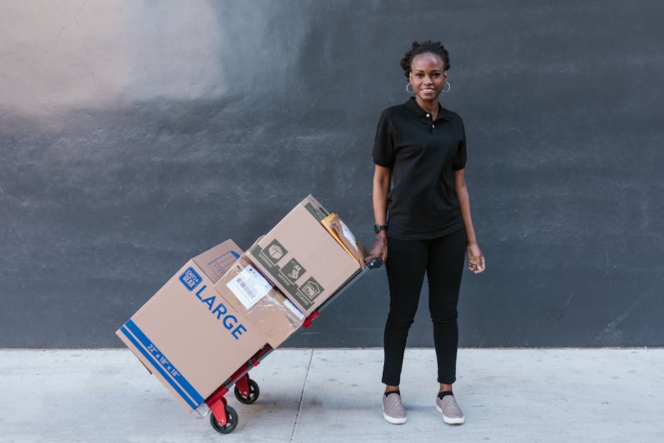Black woman in uniform pulling a dolly with large boxes, photographed outdoors against a dark wall.