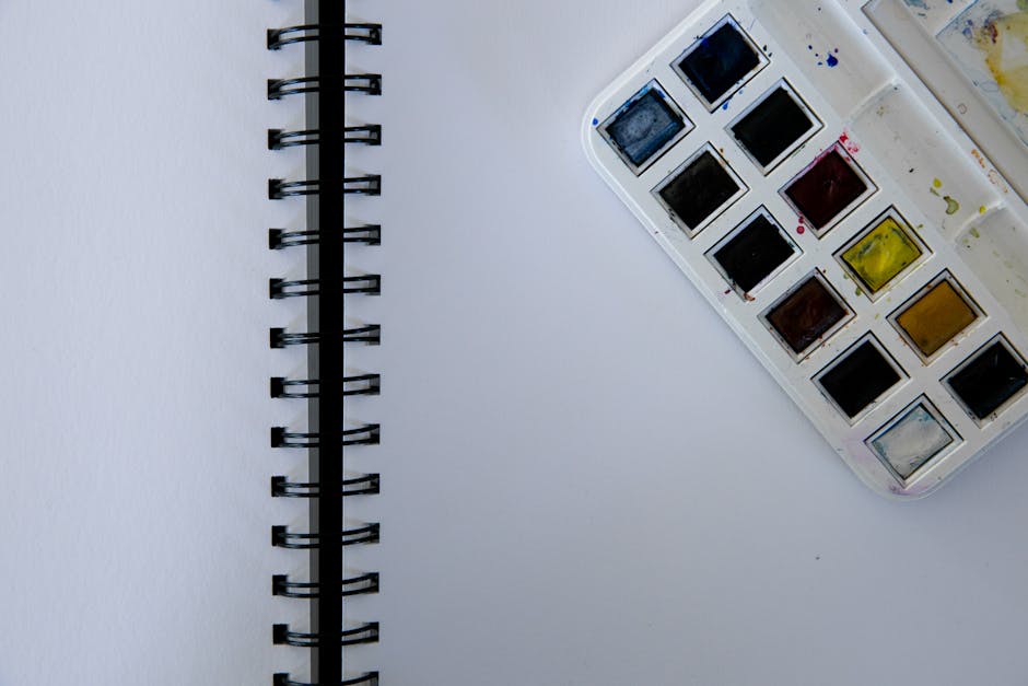 Top-down view of an open spiral notebook beside a watercolor paint set on a white background.